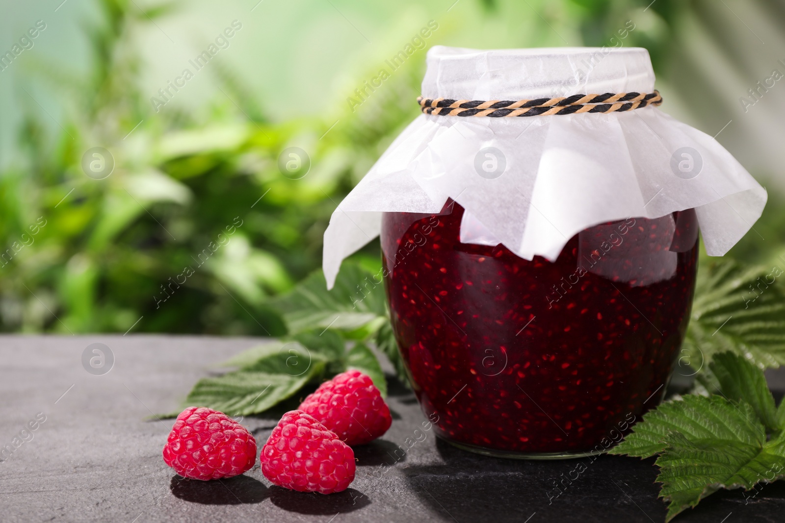 Photo of Delicious jam in glass jar and fresh raspberries on grey table outdoors. Space for text