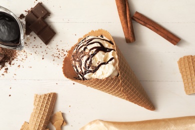 Delicious vanilla ice cream with toppings in wafer cone on white wooden table, flat lay