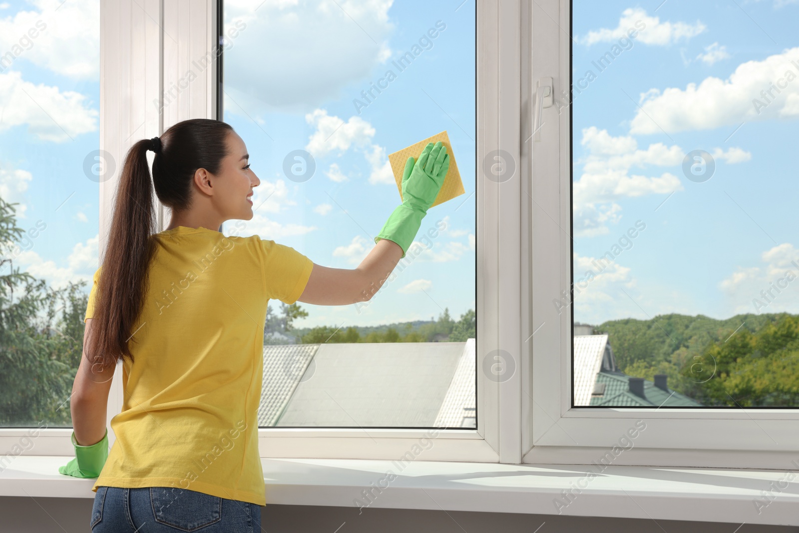 Photo of Happy young woman cleaning window glass with sponge cloth indoors