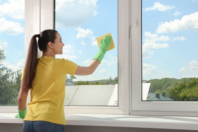 Photo of Happy young woman cleaning window glass with sponge cloth indoors