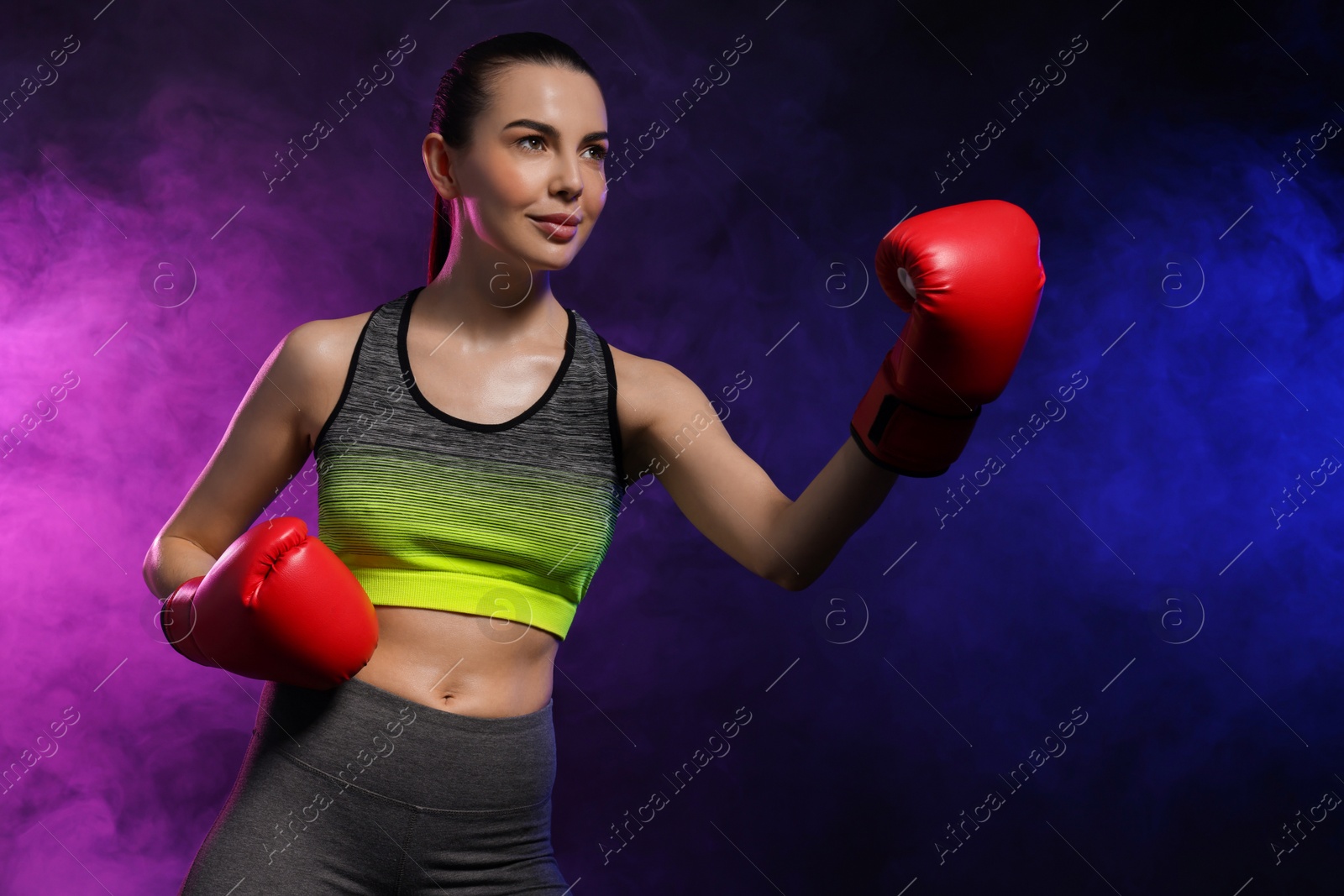 Photo of Portrait of beautiful woman wearing boxing gloves training in color lights and smoke on black background