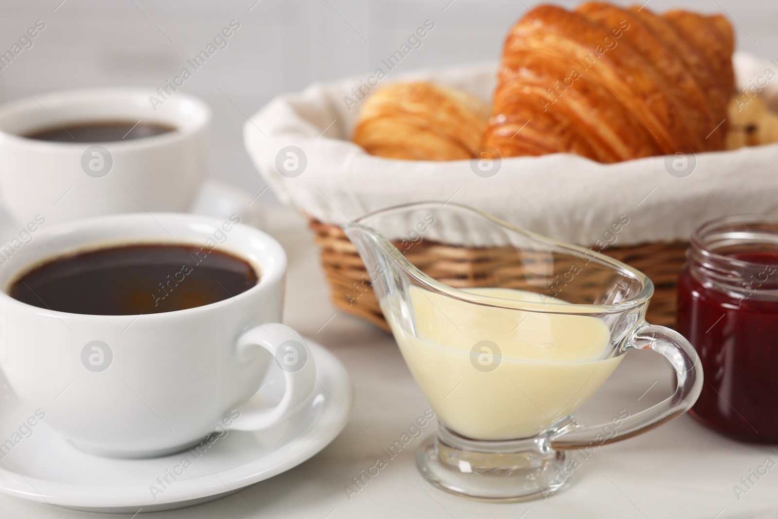 Photo of Breakfast time. Fresh croissants, coffee, sweetened condensed milk and jam on white table, closeup