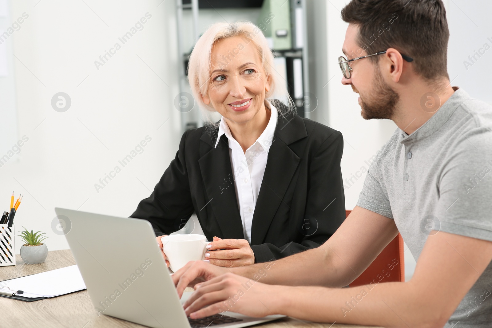 Photo of Happy boss with cup of drink and employee discussing work issues at wooden table in office