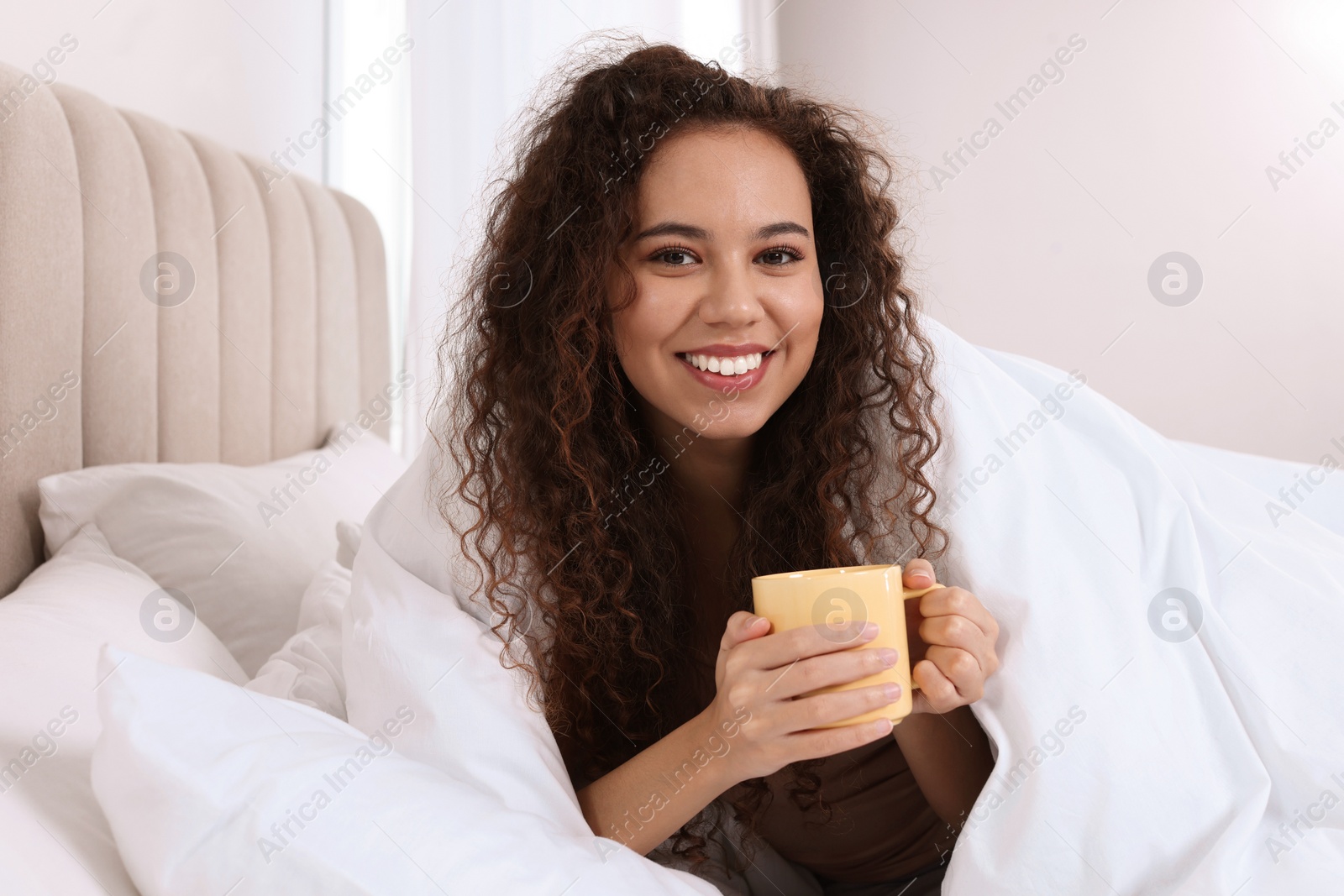 Photo of Happy African American woman with cup of drink in bed at home