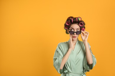 Emotional young woman in silk bathrobe with hair curlers and sunglasses on yellow background. Space for text