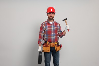 Professional builder in hard hat with hammer and tool belt on light background