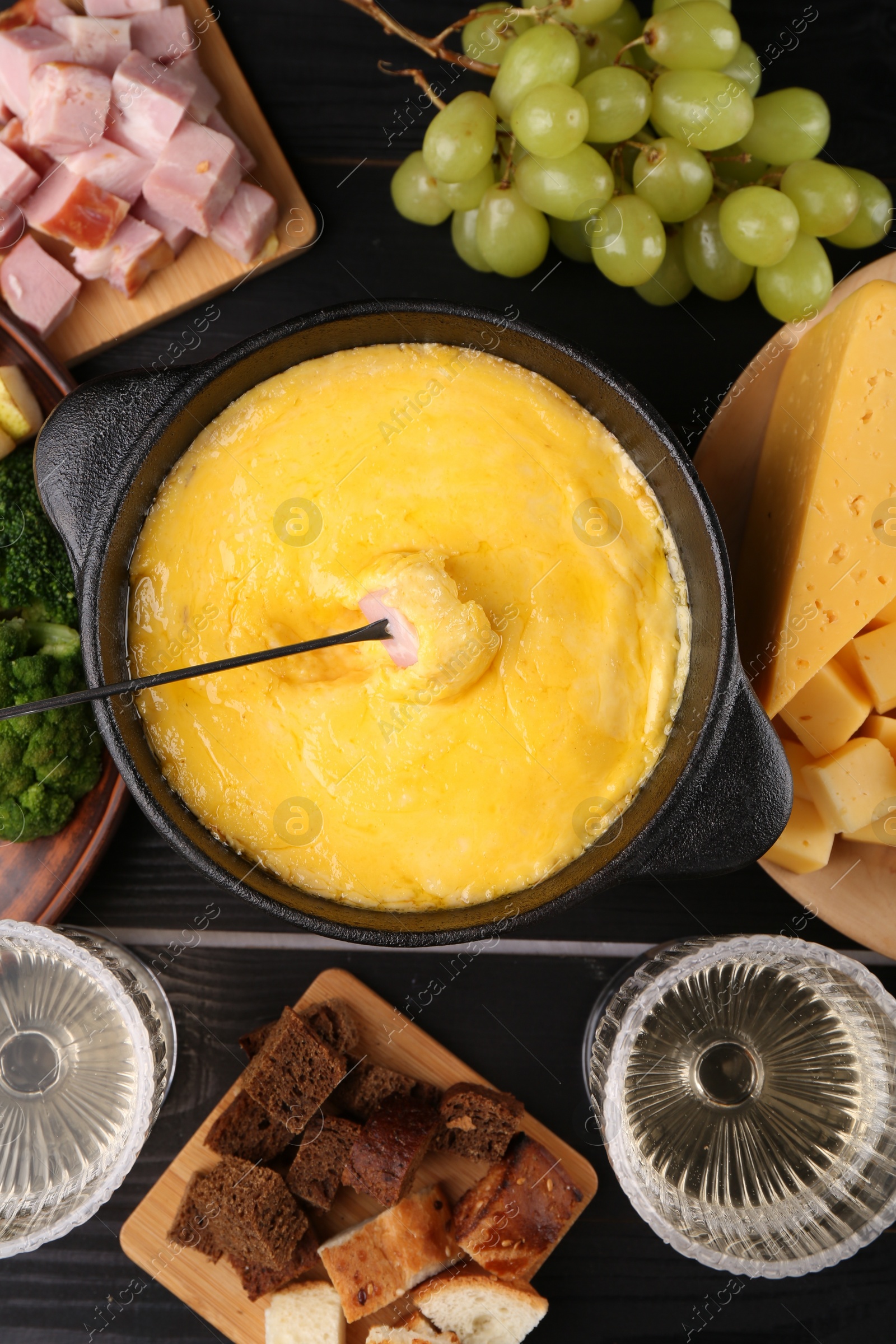 Photo of Fondue pot with melted cheese, fork and different products on black wooden table, flat lay