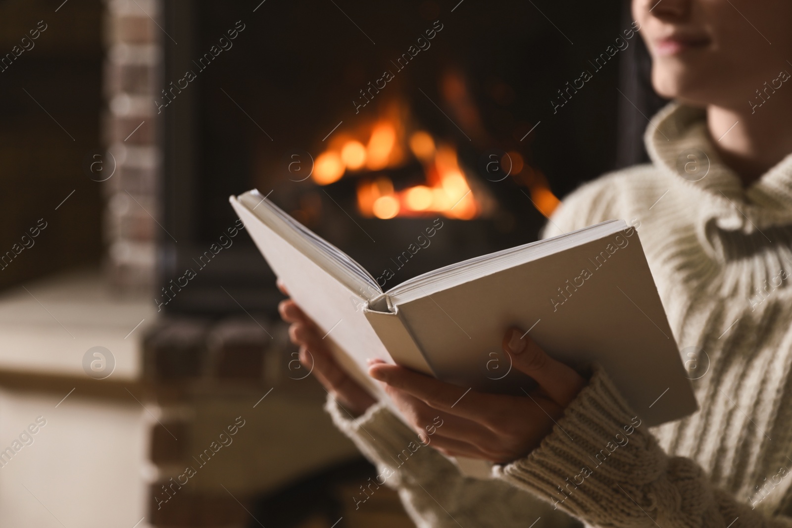 Photo of Woman reading book near burning fireplace at home, closeup