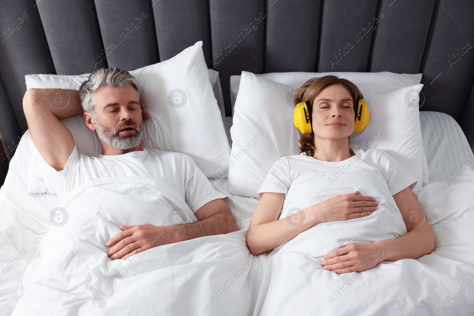 Photo of Smiling woman with headphones lying near her snoring husband in bed at home