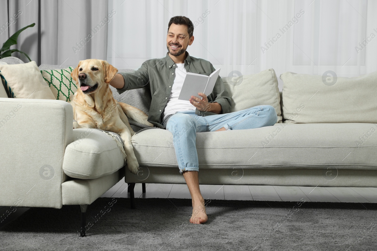 Photo of Man reading book on sofa near his cute Labrador Retriever at home