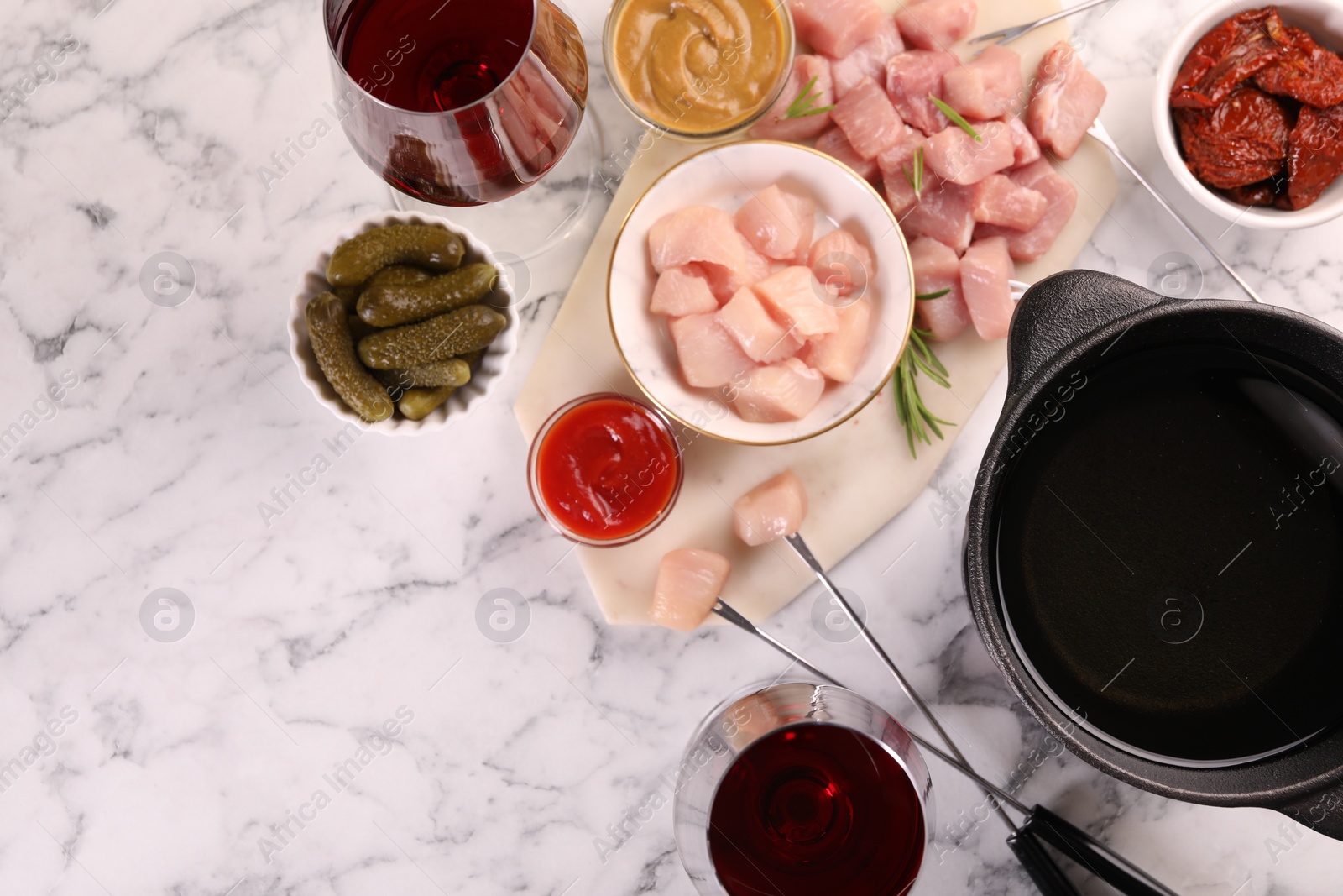 Photo of Fondue pot with oil, forks, raw meat pieces, glasses of red wine and other products on white marble table, flat lay. Space for text