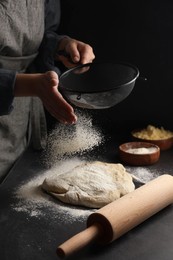 Photo of Woman sprinkling flour over dough at black table, closeup