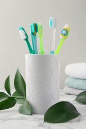 Photo of Colorful plastic toothbrushes in container and green leaves on white marble table