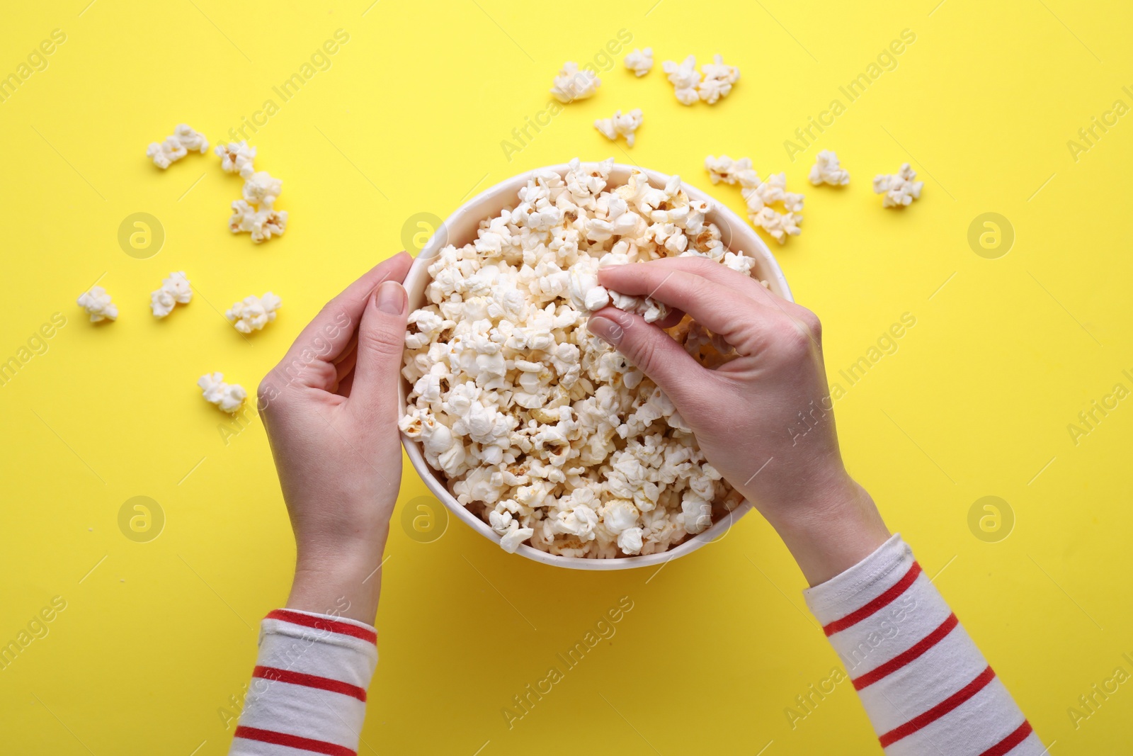 Photo of Woman taking delicious popcorn from paper bucket on yellow background, top view