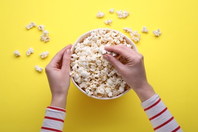 Woman taking delicious popcorn from paper bucket on yellow background, top view