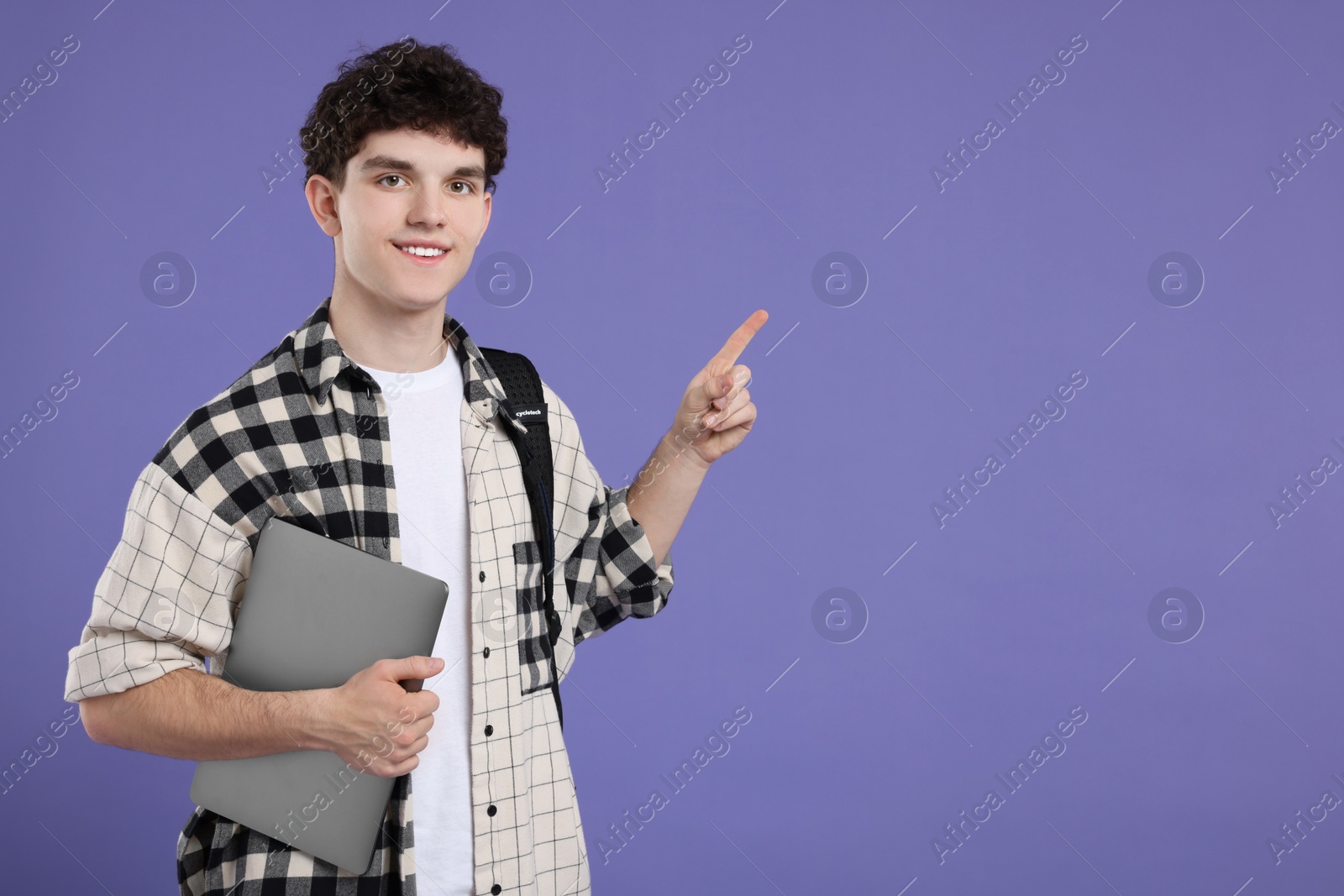 Photo of Portrait of student with backpack and laptop pointing on purple background. Space for text