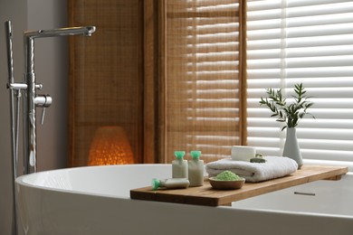 Photo of Wooden tray with spa products and green branches on bath tub in bathroom