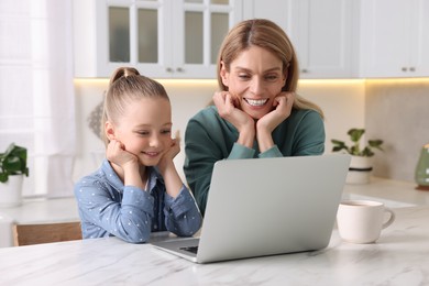Happy woman and her daughter with laptop at white table indoors