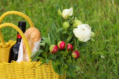 Photo of Yellow wicker bag with wine, bread and flowers on green grass outdoors, closeup. Picnic season