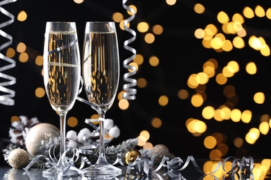 Photo of Glasses of champagne, Christmas decor and serpentine streamers against black background with blurred lights. Space for text