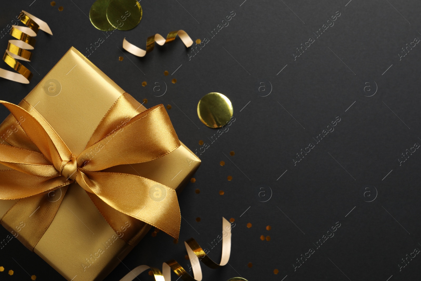 Photo of Golden gift box with confetti and streamers on black background, flat lay. Space for text