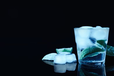 Shot glass of vodka with ice cubes and lime slices on dark background, space for text