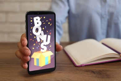 Image of Bonus gaining. Woman showing smartphone at wooden table, closeup. Illustration of open gift box, word and confetti on device screen