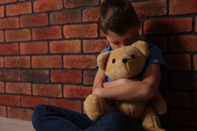 Child abuse. Upset boy with teddy bear near brick wall, space for text