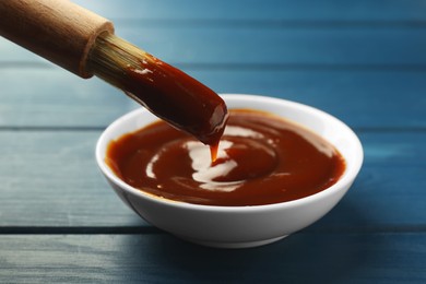 Photo of Brush with tasty barbeque sauce above bowl on blue wooden table, closeup