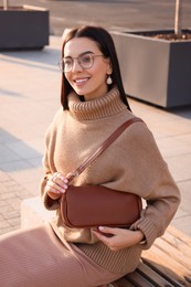 Fashionable young woman with stylish bag on bench outdoors