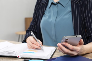 Photo of Woman taking notes while using smartphone at wooden table indoors, closeup. Space for text