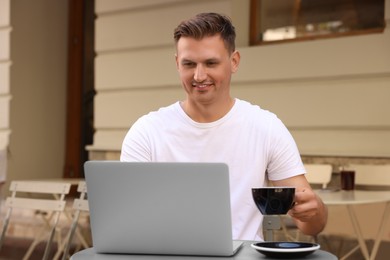 Photo of Handsome man with cup of drink working on laptop at table in outdoor cafe