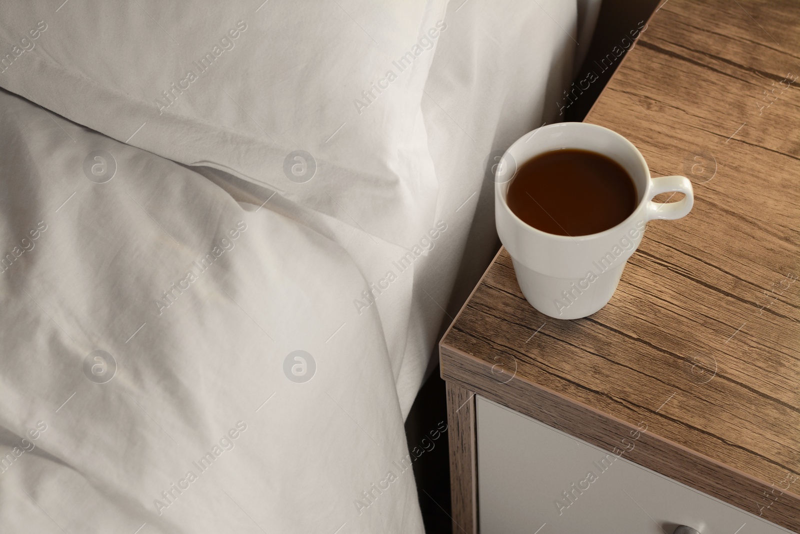 Photo of Cup of morning coffee on wooden night stand near bed indoors