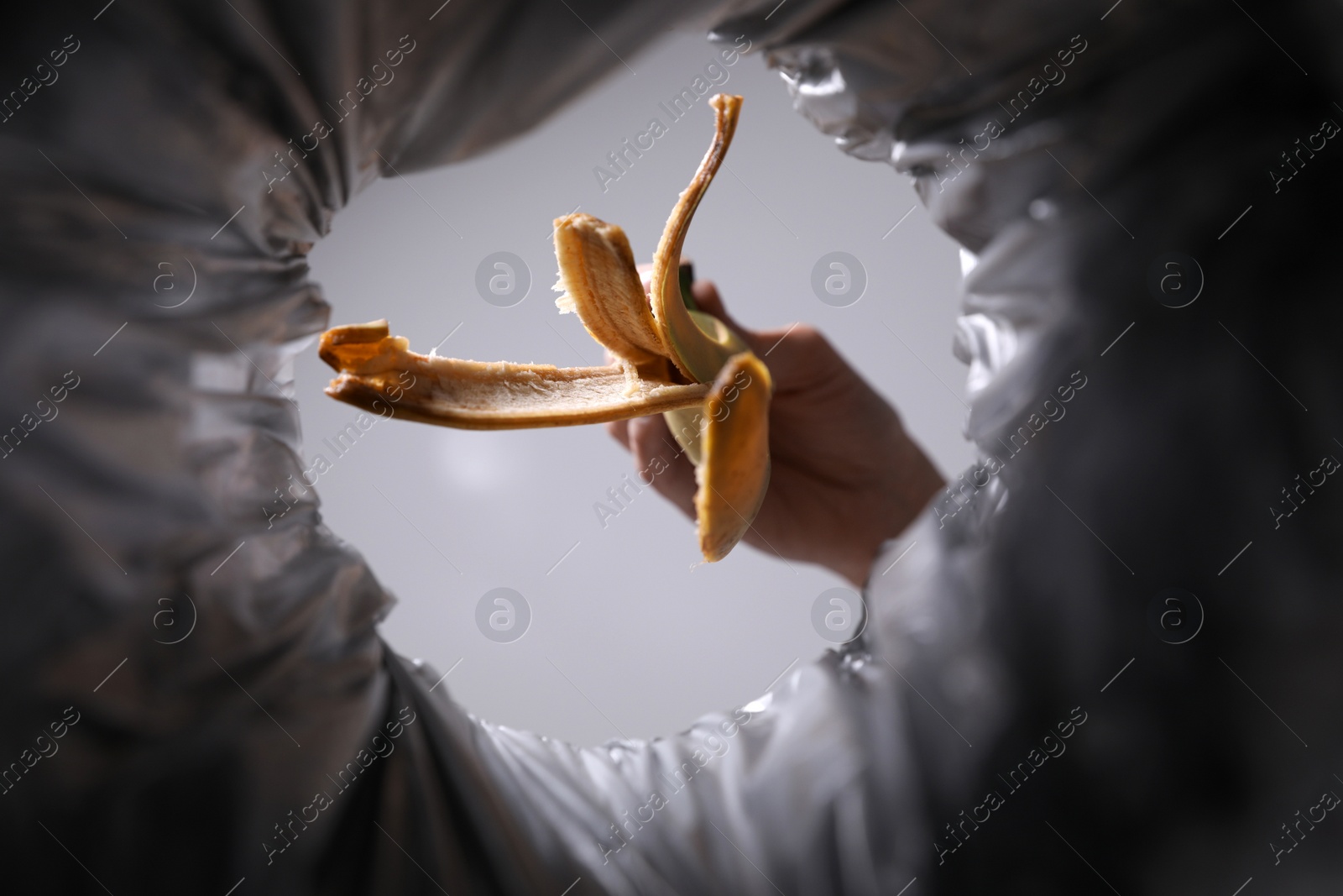 Photo of Bottom view of woman throwing banana peel into trash bin on grey background, closeup