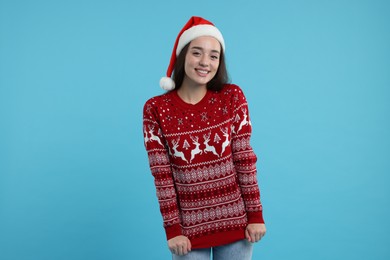 Happy young woman in Santa hat showing Christmas sweater on light blue background