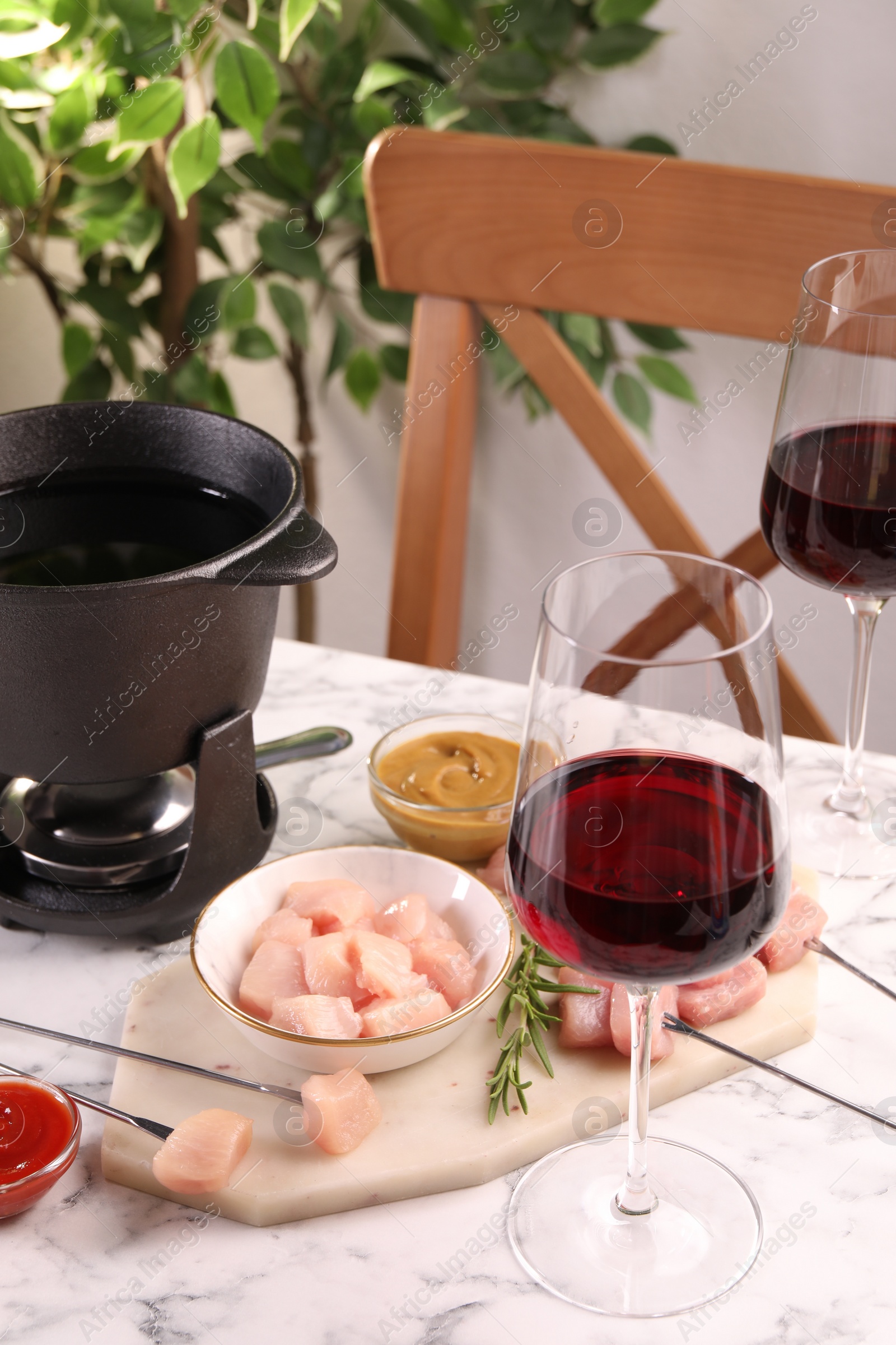 Photo of Fondue pot with oil, forks, raw meat pieces, glasses of red wine and other products on white marble table