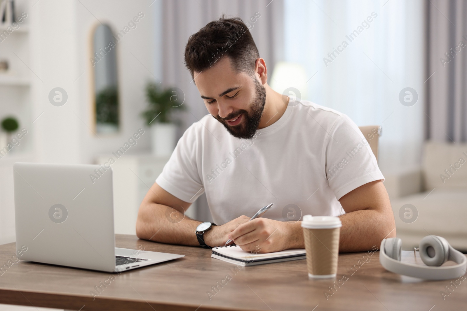 Photo of Young man writing down notes during webinar at table in room