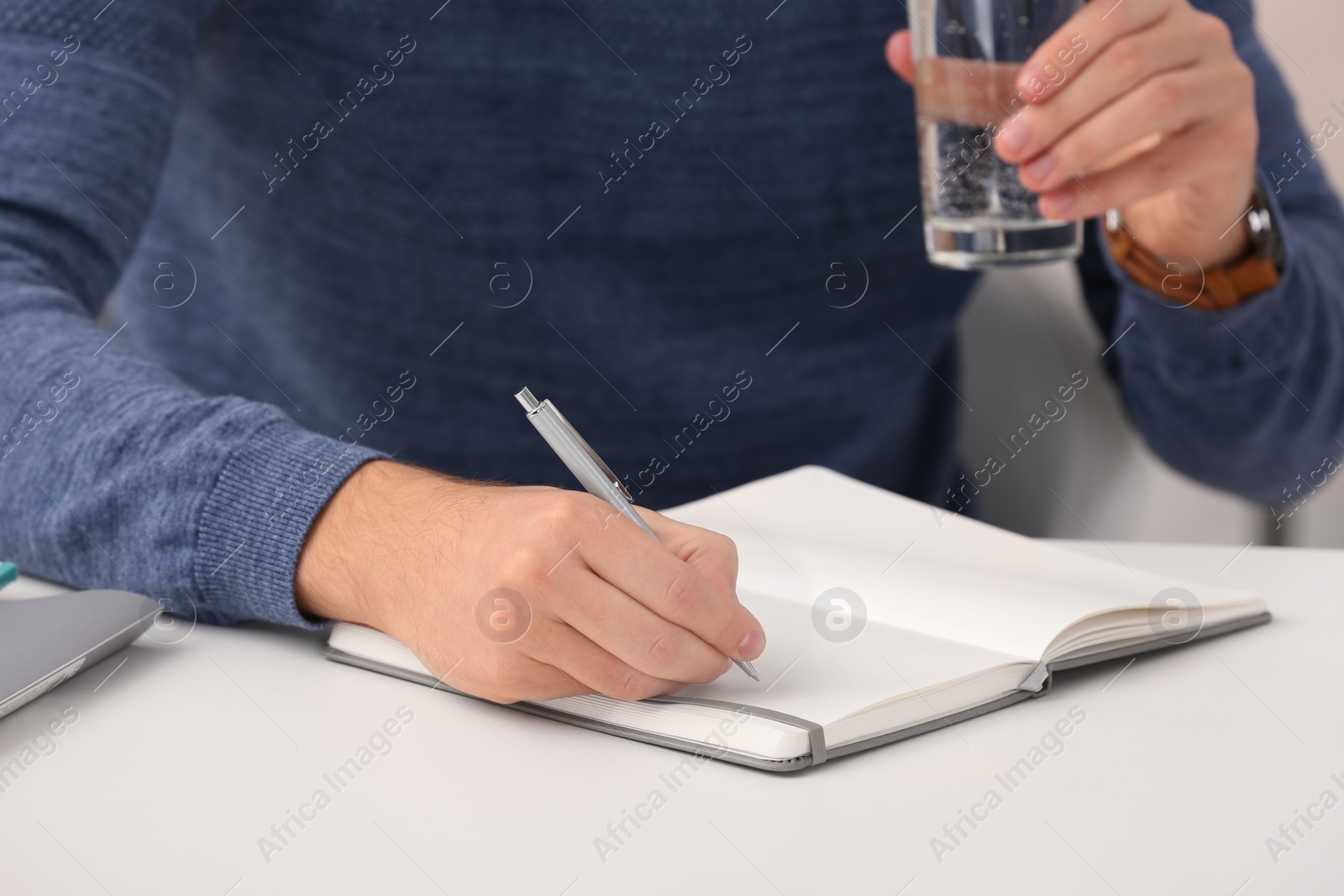Photo of Man with glass of water writing in notebook at table indoors, closeup