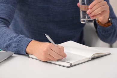 Photo of Man with glass of water writing in notebook at table indoors, closeup