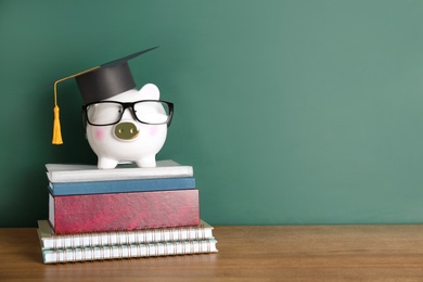 Piggy bank with graduation hat and books on table near chalkboard. Space for text