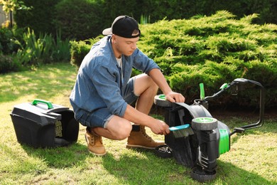 Photo of Young man cleaning lawn mower with brush in garden