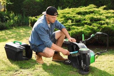 Young man cleaning lawn mower with brush in garden