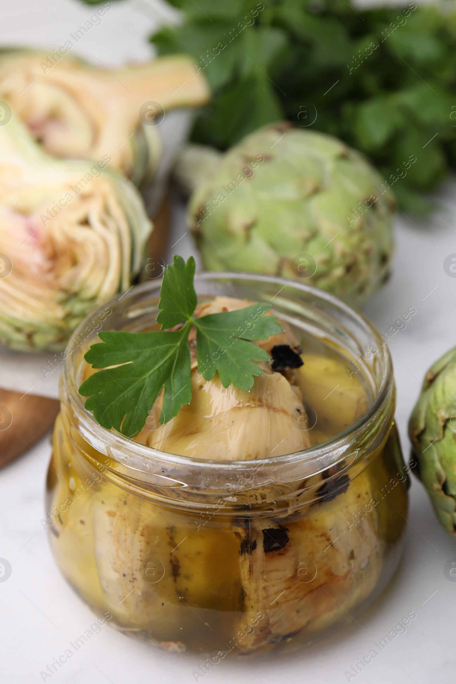 Photo of Jar of delicious artichokes pickled in olive oil and fresh vegetables on white table, closeup