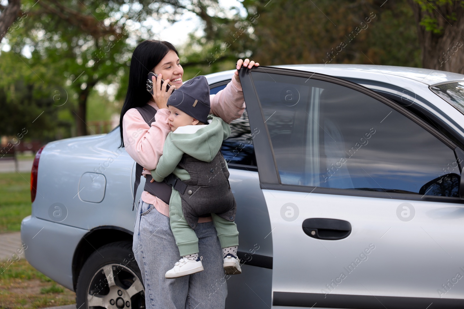 Photo of Mother holding her child in sling (baby carrier) while talking on smartphone near car outdoors