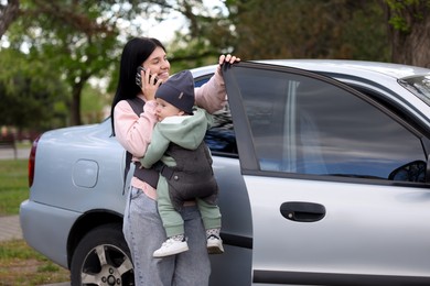 Mother holding her child in sling (baby carrier) while talking on smartphone near car outdoors