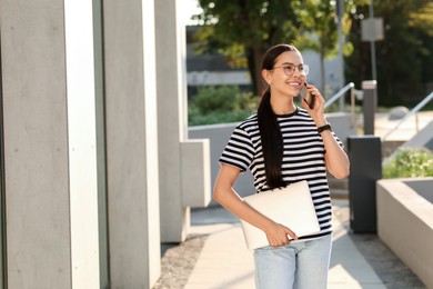 Photo of Happy young woman with modern laptop and talking on smartphone outdoors. Space for text