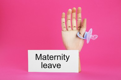 Photo of Wooden mannequin hand, baby pacifier and note with text Maternity Leave on pink background
