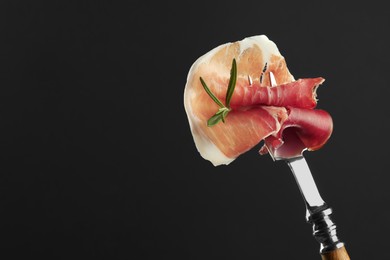 Fork with slice of tasty jamon and rosemary on black background, closeup. Space for text
