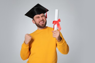 Emotional student with graduation hat and diploma on light grey background