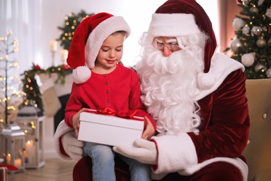 Santa Claus giving present to little girl in room decorated for Christmas